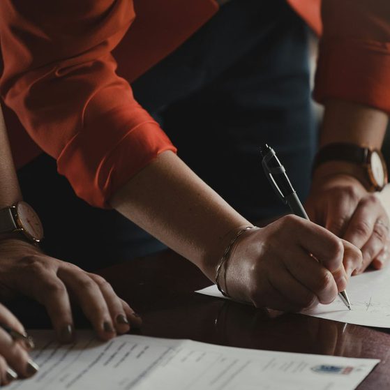 A person in an orange shirt is leaning over a table, signing a document with a pen, with their left hand resting on another document.