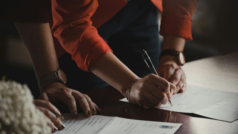 A person in an orange shirt is leaning over a table, signing a document with a pen, with their left hand resting on another document.