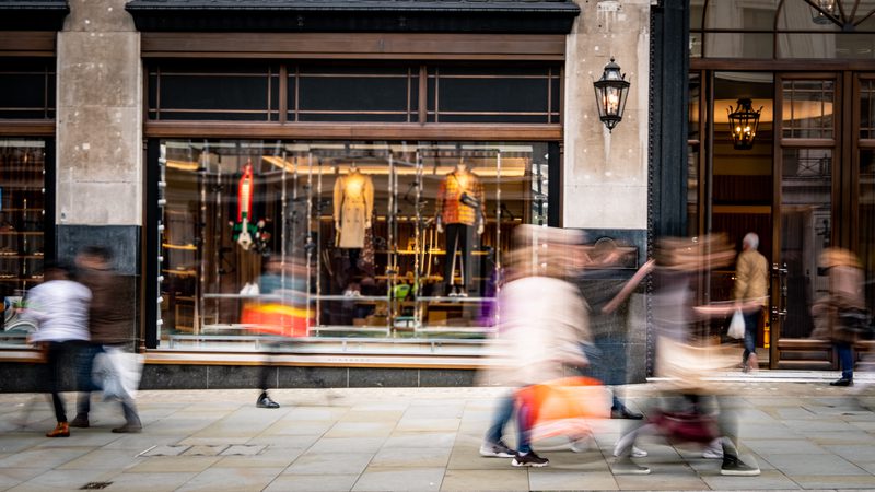 Blurred pedestrians walking past a clothing store window display on a city street with stone pavement. The store features mannequins in jackets and scarves inside.
