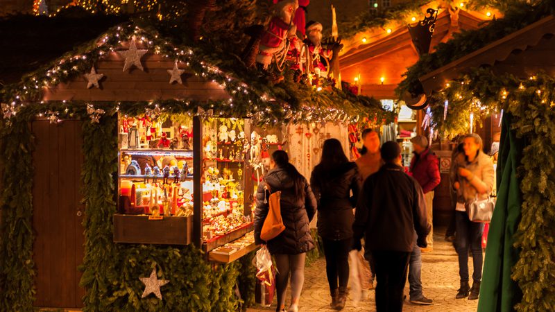 A warmly lit, festive Christmas market with decorated wooden stalls, shoppers browsing ornaments and gifts, and holiday greenery and lights creating a cozy atmosphere.
