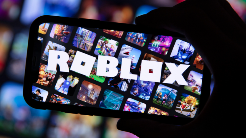 A hand holds a smartphone displaying the Roblox logo amid a grid of colourful game thumbnails on a vibrant, blurred background with small square Roblox icons filling the screen.