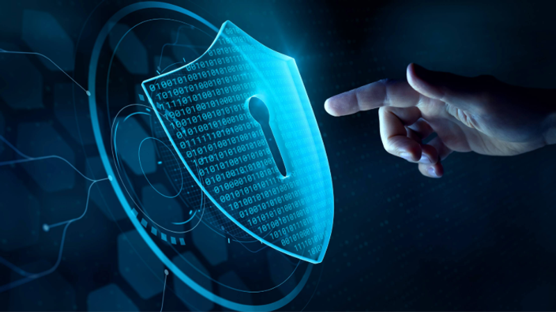 A blue holographic shield with a binary code pattern and a central keyhole glows from a digital panel as a finger points toward it, symbolising cybersecurity.