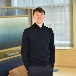 A young man with short brown hair wearing a black zip-up sweater and grey trousers, standing indoors with hands in pockets and smiling.