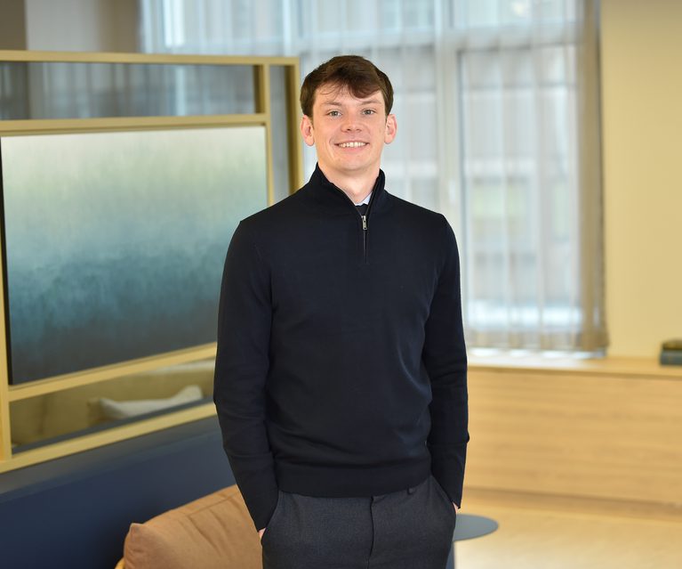 A young man with short brown hair wearing a black zip-up sweater and grey trousers, standing indoors with hands in pockets and smiling.
