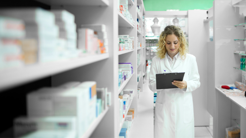 A person in a white lab coat stands in a bright pharmacy aisle, holding a clipboard and checking notes beside shelves of medicines.