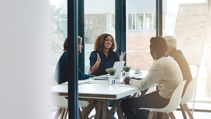 A diverse group of four people having a discussion around a white conference table in a bright, modern office with large windows and a whiteboard in the background.