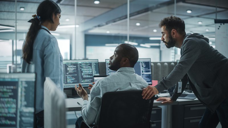 Three individuals are engaged in a discussion in a modern office with multiple computer monitors displaying code. One person is seated, while the other two stand and converse with him.