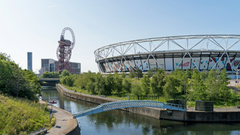 A calm canal with a pale-blue arched footbridge, lush trees and West Ham's London stadium in the background.