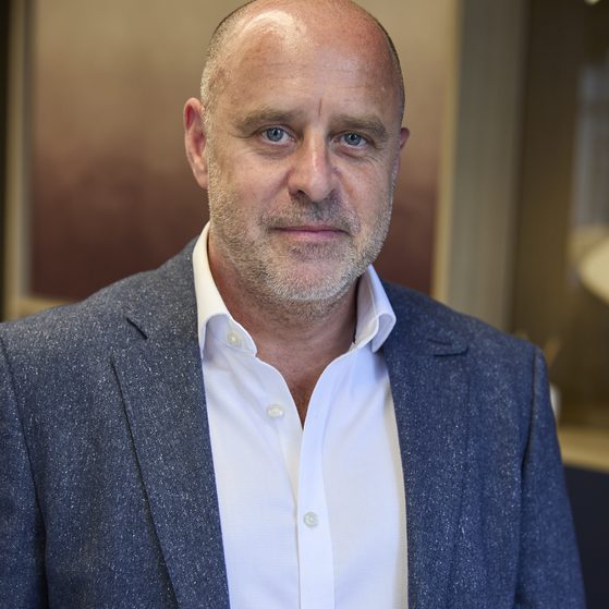 Adult with a shaved head and light stubble wearing a white shirt and blue speckled blazer, posed in a warmly lit indoor setting.