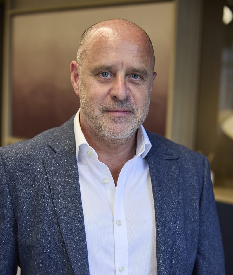 Adult with a shaved head and light stubble wearing a white shirt and blue speckled blazer, posed in a warmly lit indoor setting.