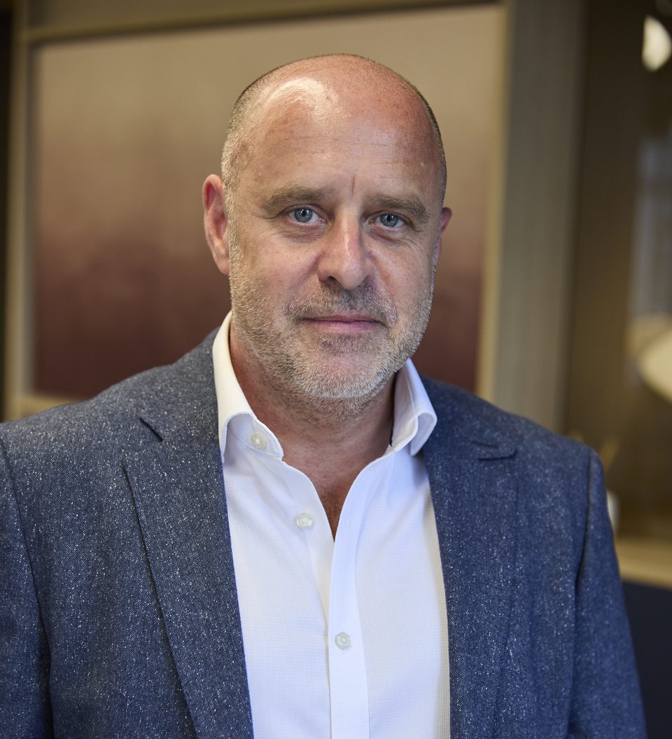 Adult with a shaved head and light stubble wearing a white shirt and blue speckled blazer, posed in a warmly lit indoor setting.