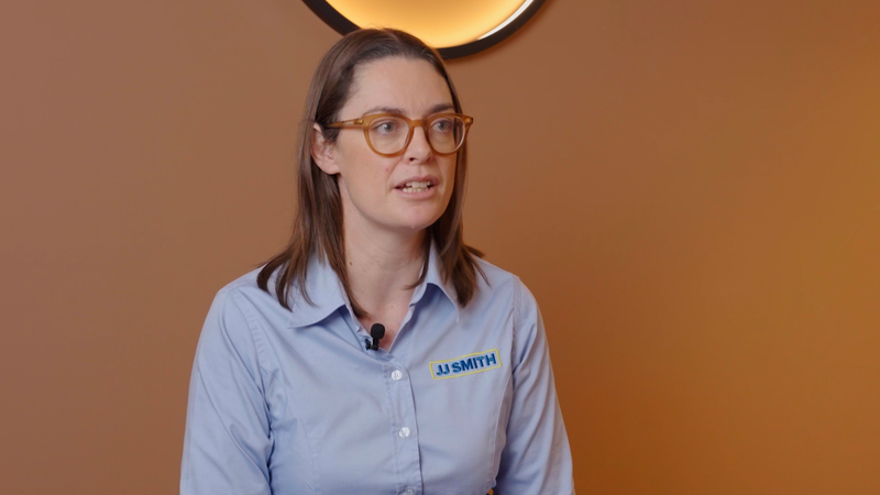 A woman with brown hair and glasses wearing a light blue shirt branded "JJ SMITH" in front of a warm orange wall, speaking into a microphone.