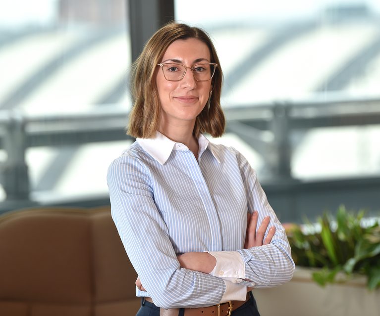 A woman with shoulder-length brown hair, wearing glasses and a light blue striped shirt, stands confidently with arms crossed in a modern office with large windows and plants.