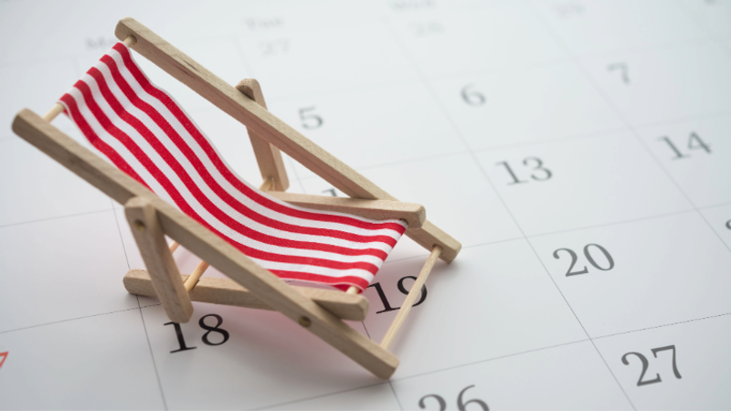 A small wooden beach chair with red and white striped fabric sits on a calendar page, suggesting summer holidays and planning for upcoming dates.