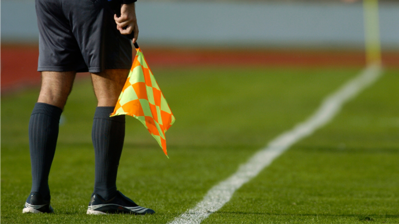Lower half of a football assistant linesman on the pitch, wearing dark shorts and socks, holding a bright orange and yellow checkered flag beside a chalked sideline.
