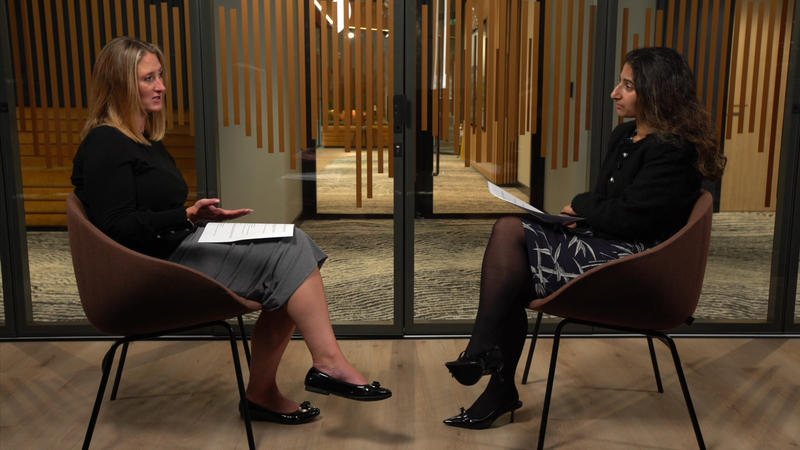 Two women are seated in chairs facing each other, having a conversation in a modern office setting with wooden decor and glass partitions.