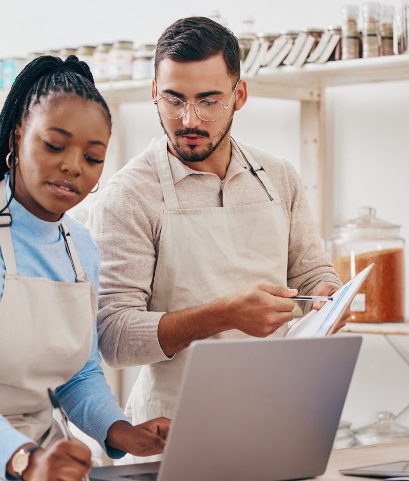 A man and woman wearing aprons are working together in a shop, looking at a laptop and a notebook in a well-lit space with shelves of jars and products behind them.