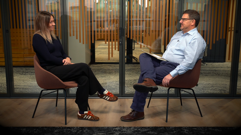 A woman and a man are sitting in chairs facing each other, engaged in a conversation in a modern office setting with glass walls and wooden accents.