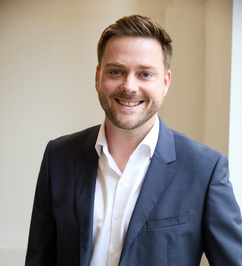 A person with short brown hair, blue eyes and a trimmed beard wears a navy suit over a white shirt, smiling at the camera indoors beside a light wall.