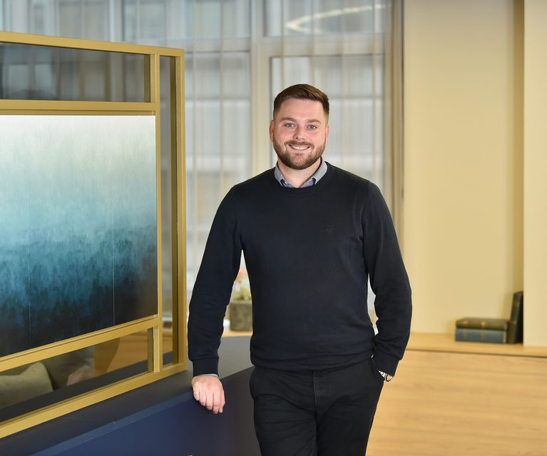 A man with short brown hair and a beard, wearing a dark sweater over a collared shirt, stands smiling in a modern office space with large windows.