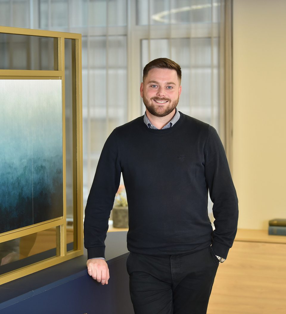A man with short brown hair and a beard, wearing a dark sweater over a collared shirt, stands smiling in a modern office space with large windows.