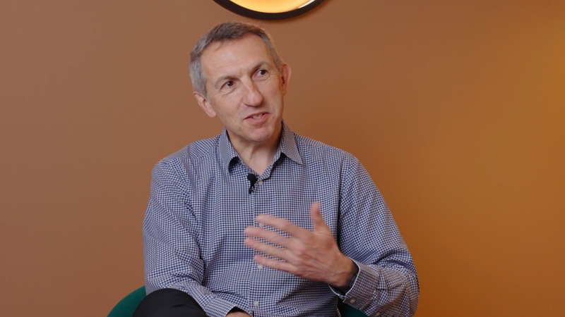 A middle-aged man with short grey hair, wearing a checkered shirt, is sitting against an orange wall with a circular wall clock behind him, gesturing with his right hand on his chest.