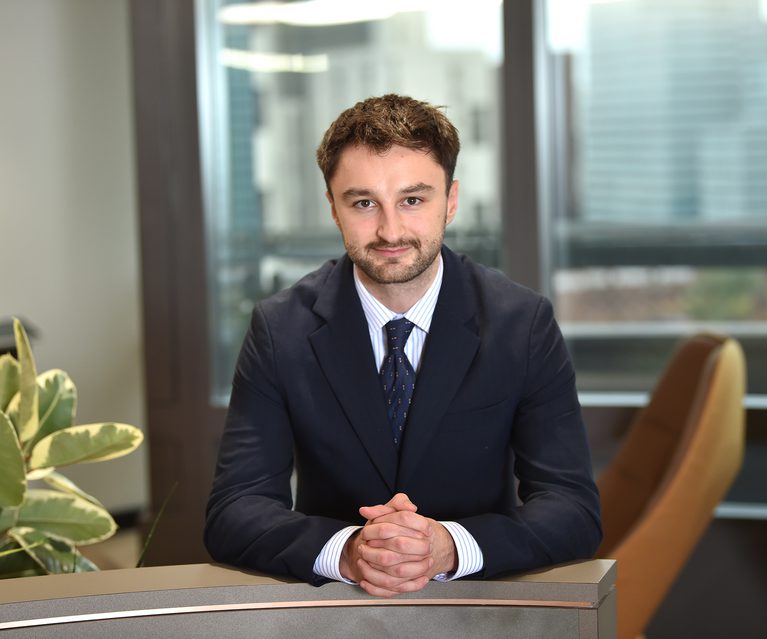 A young man with brown hair and a beard, wearing a dark suit and striped shirt, sitting at a desk with hands clasped, in an office with large windows.