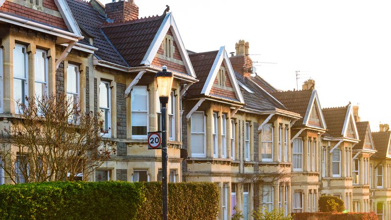 Row of Victorian-style terraced houses with bay windows, brick and stone facades, and pitched roofs, illuminated by warm sunlight.
