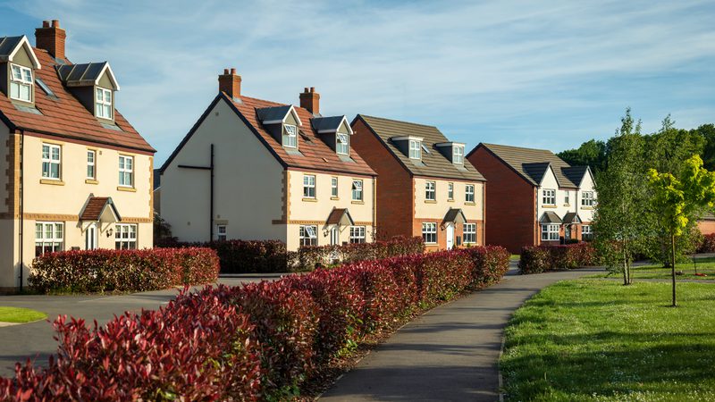 Row of modern houses with red brick and white exteriors, surrounded by colourful bushes and green lawns, under a partly cloudy sky.