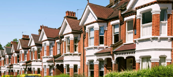 Row of red-brick terraced houses with white detailing, gabled roofs and chimneys, front gardens, and cars parked along a sunny street.