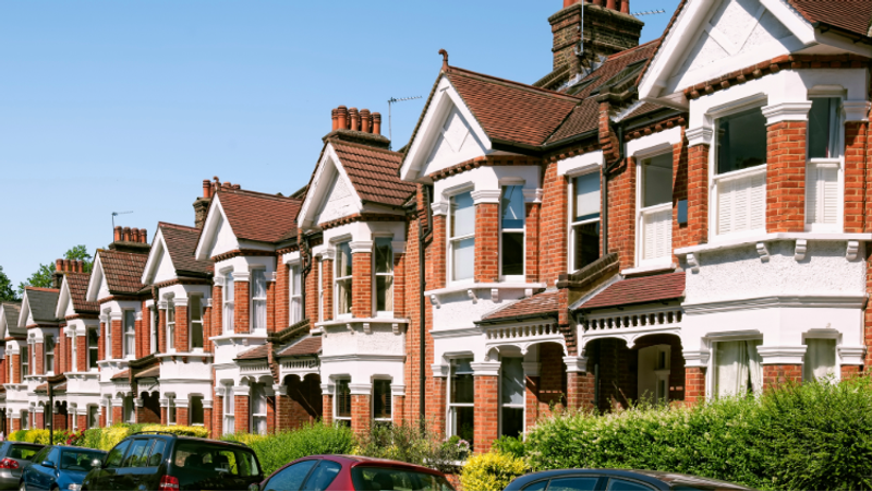 Row of red-brick terraced houses with white detailing, gabled roofs and chimneys, front gardens, and cars parked along a sunny street.