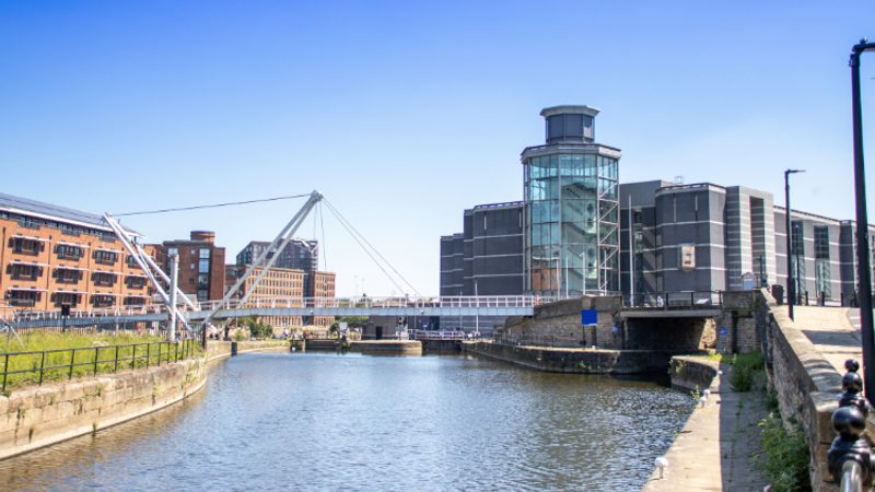 Sunny urban canal scene featuring Royal Armouries Museum and New Dock and a white cable-stayed pedestrian bridge spanning the water.
