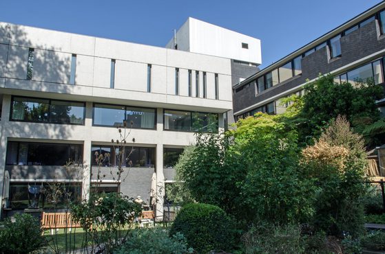 Modern apartment buildings with large windows, surrounded by lush green bushes and trees, under a clear blue sky.