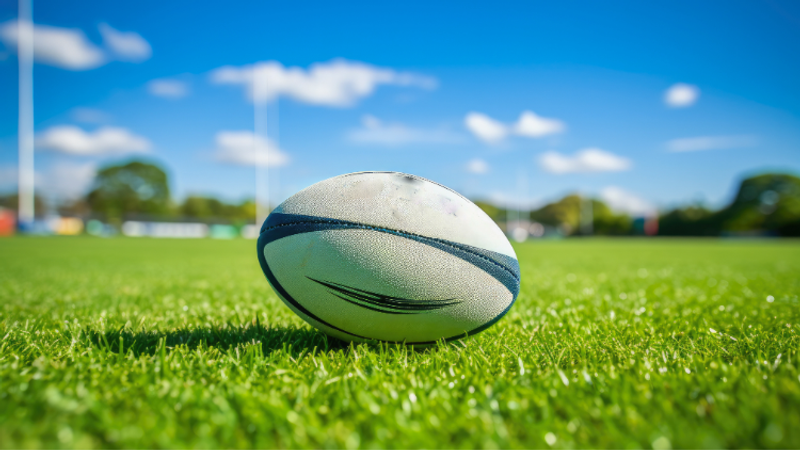 Rugby ball rests on bright green grass of an outdoor field, under a blue sky with fluffy clouds and distant goalposts in the blurred background.
