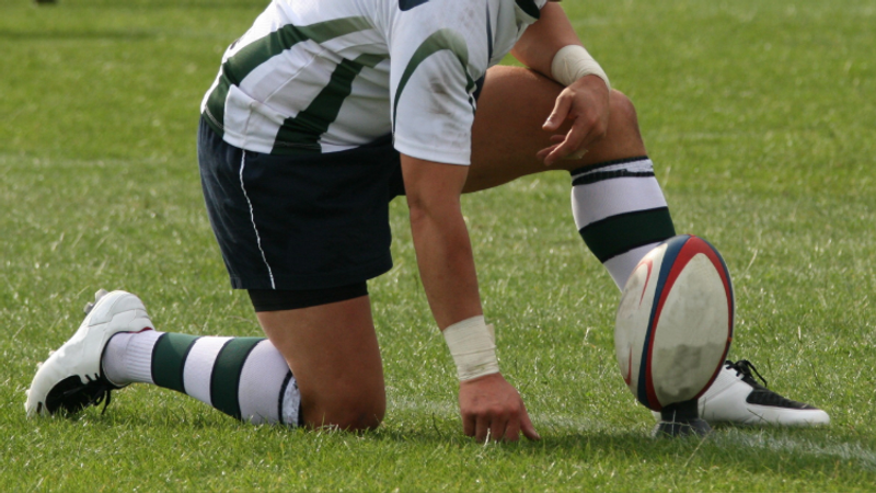 A rugby player kneeling on the grass, with one knee down and the other foot flat on the ground, holding a rugby ball in front of a green field.