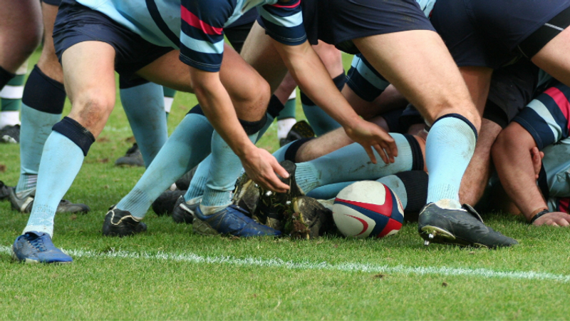 Group of rugby players forming a scrum on a grass field, with focus on their legs, hands, and rugby ball in the centre.