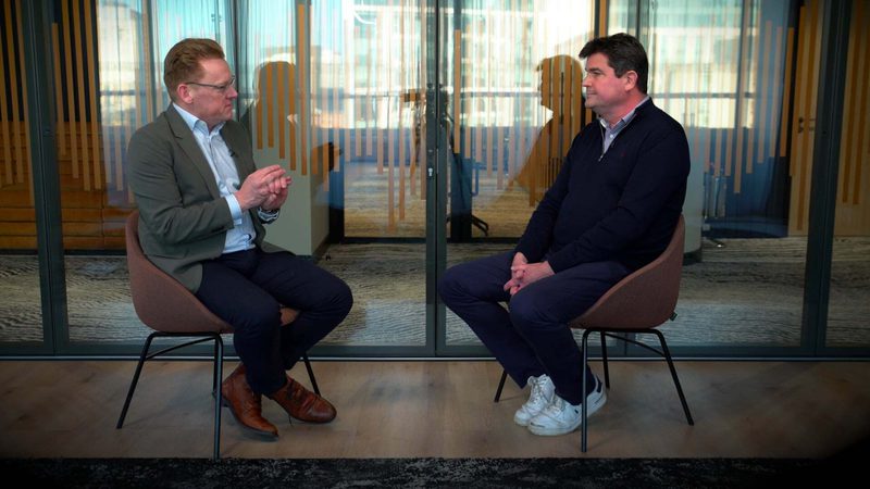 Two men sit facing each other in a modern interview setting, separated by a glass partition, with reflections of other people behind them.