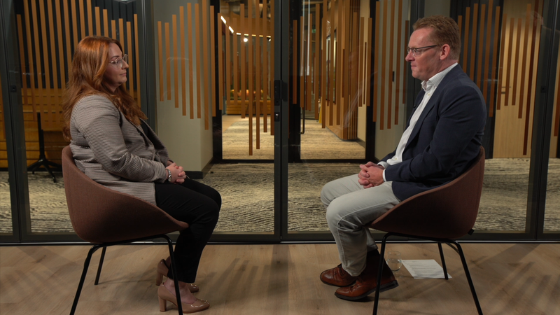 A woman with long red hair and glasses, wearing a checkered blazer, sits facing a man with glasses, in a navy blazer, grey trousers, and brown shoes; they are engaged in a conversation in a modern room with a glass wall and wooden accents.