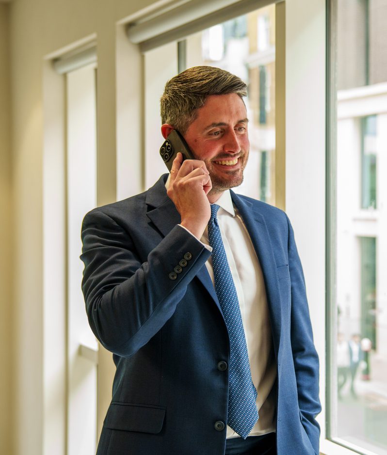 Person in a navy suit talks on a smartphone by a large window in a bright office, with tall glass buildings and a city street visible outside