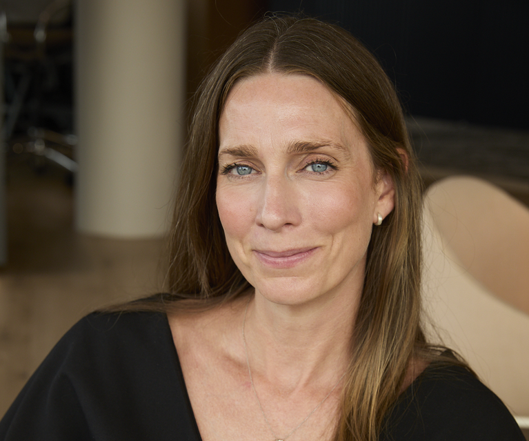 A person with shoulder-length light brown hair and blue eyes sits in a black V-neck dress with a wide belt, smiling softly in a warm indoor setting.