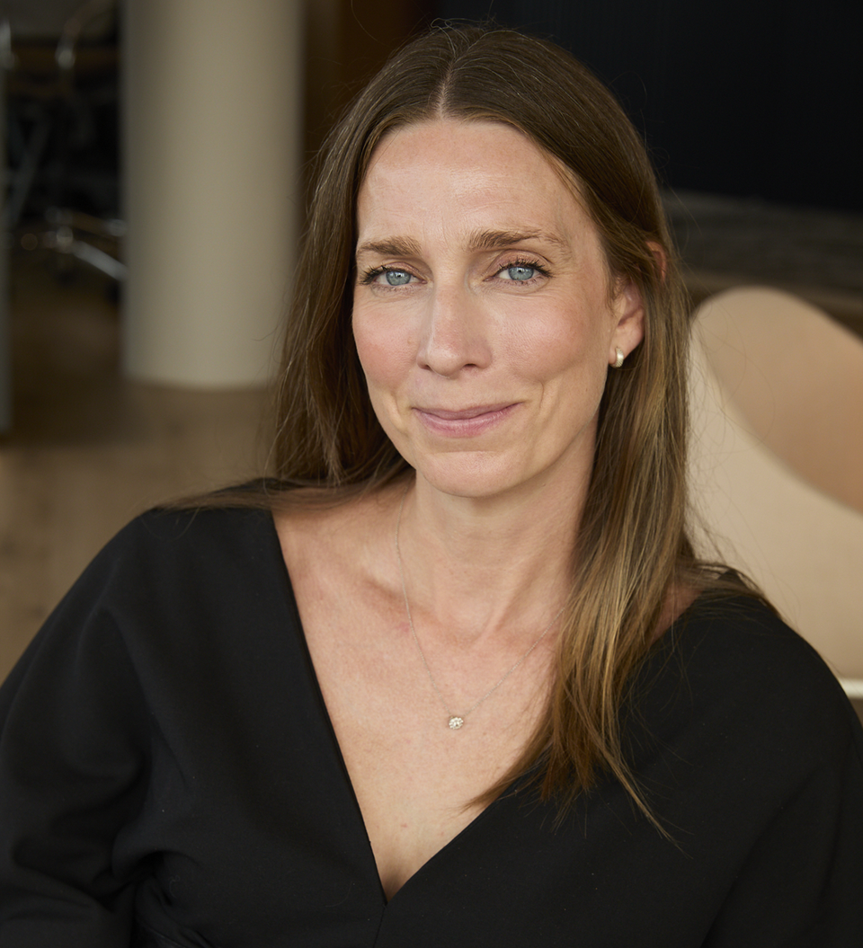 A person with shoulder-length light brown hair and blue eyes sits in a black V-neck dress with a wide belt, smiling softly in a warm indoor setting.