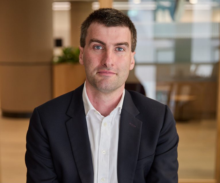 A person with short dark hair wearing a navy blazer and white shirt sits in a modern office lobby, with glass partitions and warm lighting in the background.