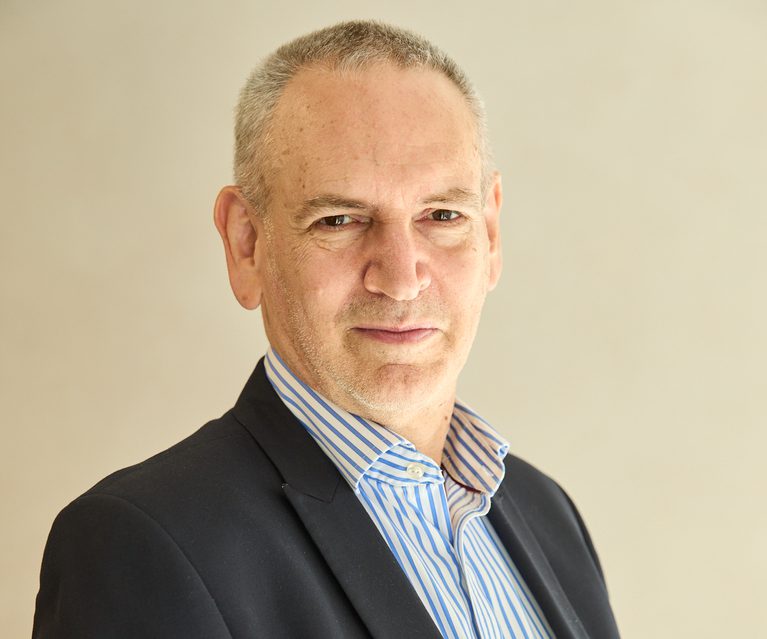 Head-and-shoulders portrait of a person with short grey hair, wearing a dark blazer and blue striped shirt, against a plain beige background.