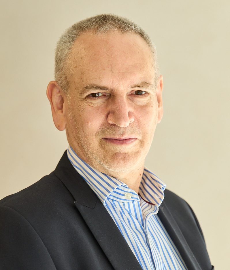 Head-and-shoulders portrait of a person with short grey hair, wearing a dark blazer and blue striped shirt, against a plain beige background.