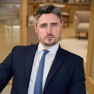 Person in a navy suit, white shirt and patterned blue tie sits in a modern office lounge, looking at the camera with a composed, serious expression.