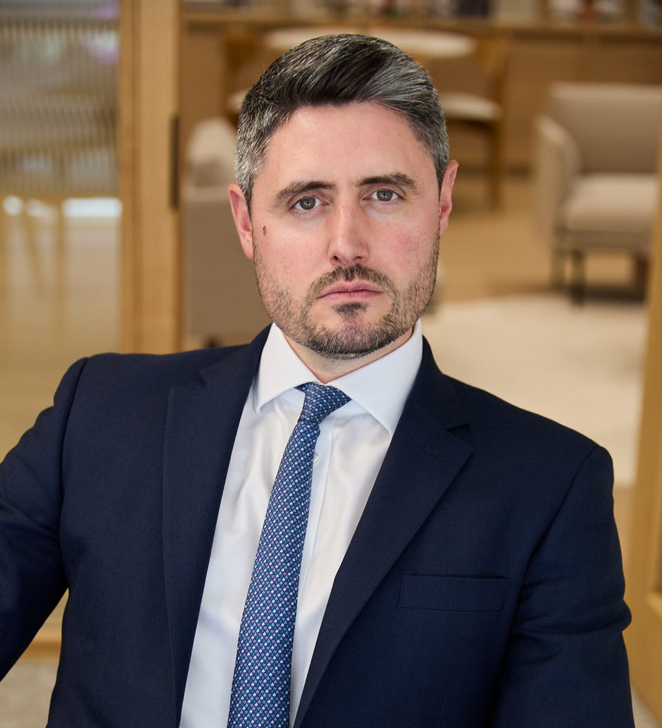 Person in a navy suit, white shirt and patterned blue tie sits in a modern office lounge, looking at the camera with a composed, serious expression.