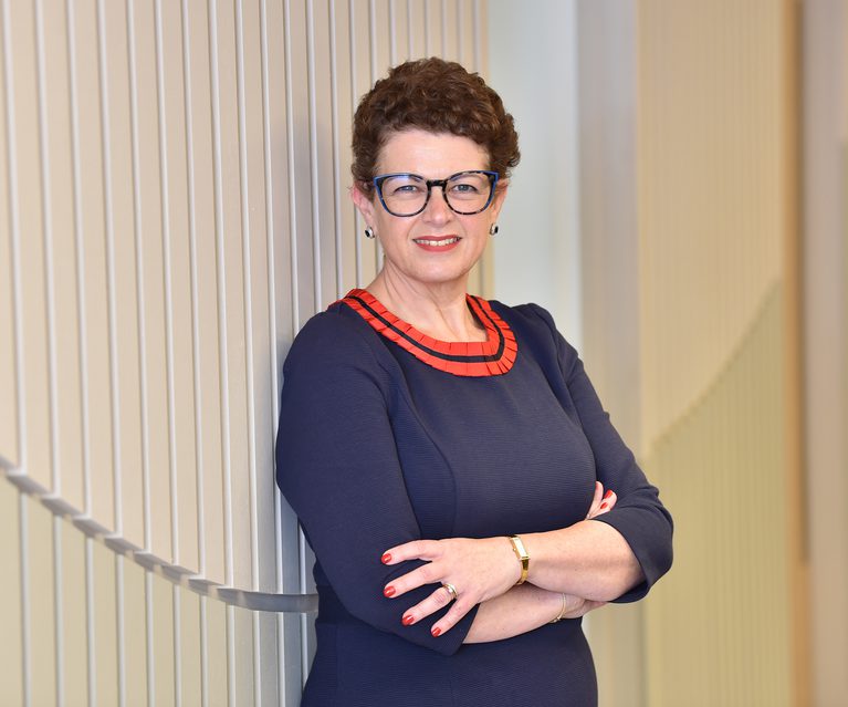 A woman with short curly brown hair, glasses, and earrings, wearing a navy blue dress with red detailing, stands with arms crossed in front of a light-colored wall.