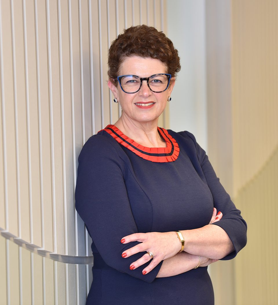 A woman with short curly brown hair, glasses, and earrings, wearing a navy blue dress with red detailing, stands with arms crossed in front of a light-colored wall.