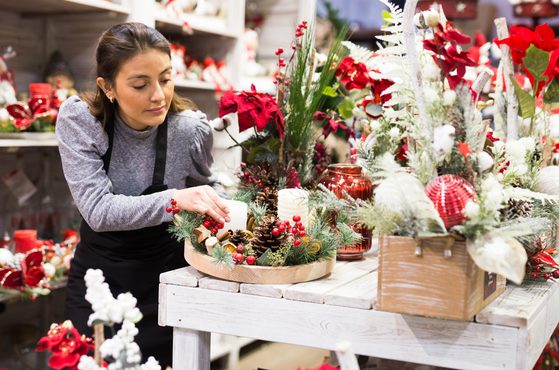 A woman arranges Christmas decorations on a white wooden table amid festive items like pine cones, berries, and red and white ornaments.