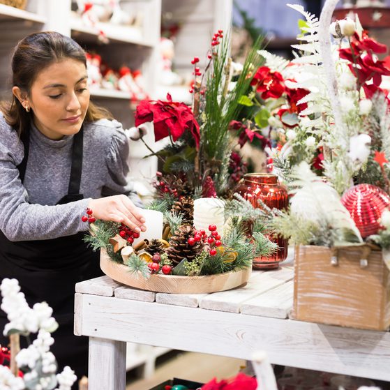 A woman arranges Christmas decorations on a white wooden table amid festive items like pine cones, berries, and red and white ornaments.